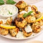 A plate of Greek chicken meatballs garnished with lemon slices and herbs, served with a small bowl of creamy dipping sauce topped with chopped parsley.