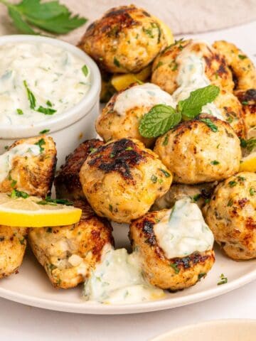 A plate of Greek chicken meatballs garnished with lemon slices and herbs, served with a small bowl of creamy dipping sauce topped with chopped parsley.