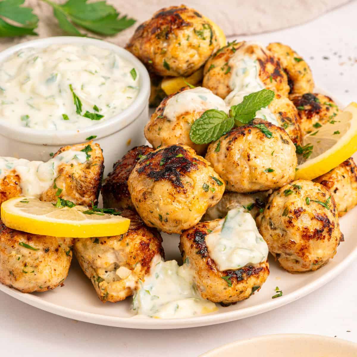 A plate of Greek chicken meatballs garnished with lemon slices and herbs, served with a small bowl of creamy dipping sauce topped with chopped parsley.