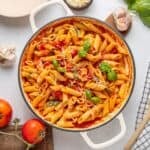 A pot of Penne Pomodoro with Italian tomato sauce, fresh basil leaves, and grated cheese, surrounded by tomatoes, garlic, basil, and a checked cloth on a white surface-a classic Italian dish.
