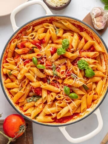 A pot of Penne Pomodoro with Italian tomato sauce, fresh basil leaves, and grated cheese, surrounded by tomatoes, garlic, basil, and a checked cloth on a white surface-a classic Italian dish.