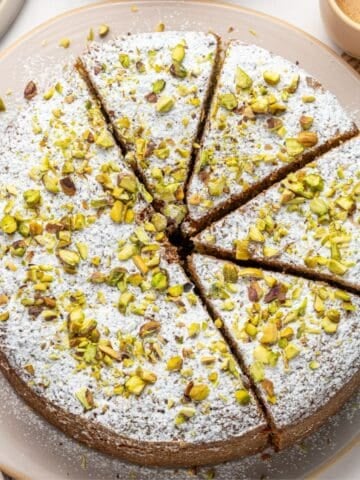 A pistachio ricotta cake, topped with powdered sugar and chopped pistachios, is cut into eight slices and displayed on a beige plate. A cup of coffee and a piece of newspaper are seen in the background.