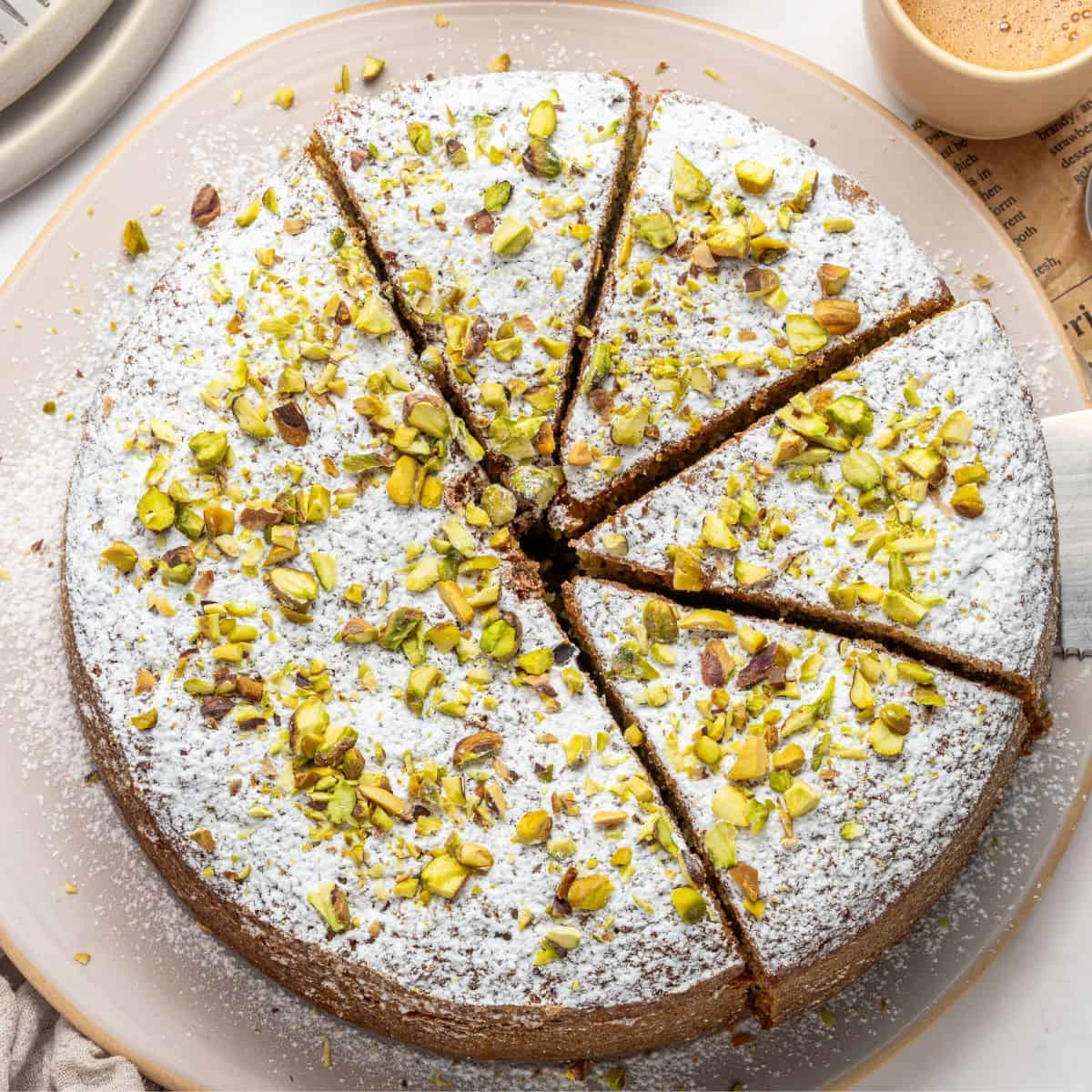 A pistachio ricotta cake, topped with powdered sugar and chopped pistachios, is cut into eight slices and displayed on a beige plate. A cup of coffee and a piece of newspaper are seen in the background.