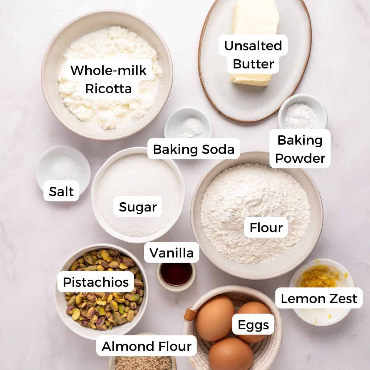 A flat lay of baking ingredients for a pistachio ricotta cake in bowls and plates, each labeled: whole-milk ricotta, unsalted butter, baking powder, baking soda, flour, lemon zest, eggs, almond flour, pistachios, vanilla, sugar, and salt.