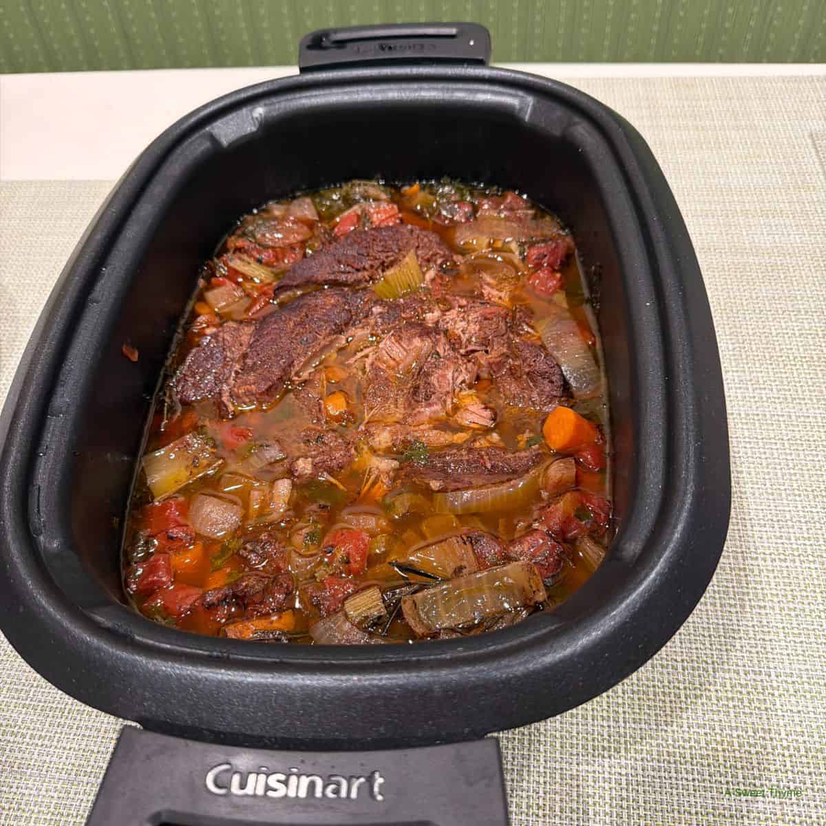 A Cuisinart slow cooker filled with pot roast—tender beef, carrots, onions, celery, and tomatoes simmer in rich broth. Perfect for a cozy night with The Sunday Thymes on your lap and comfort food on your table.