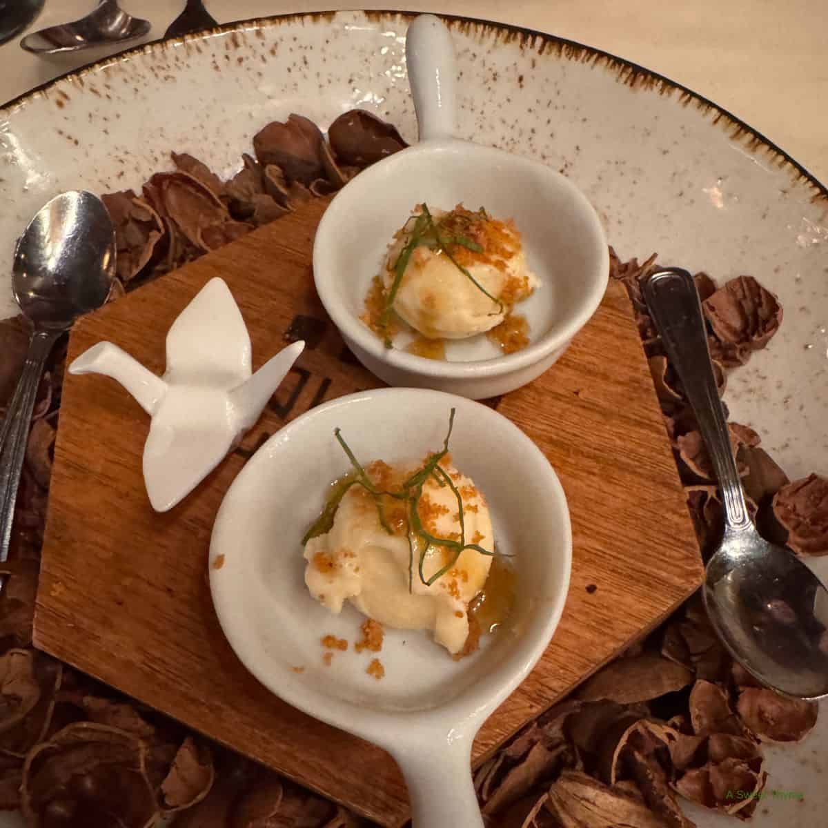 Two small white bowls with scoops of ice cream, garnished with green herbs and crumbs, are placed on a wooden board surrounded by empty nutshells—an inviting Sunday Thymes setting, complete with two spoons and a white origami-style ceramic bird.