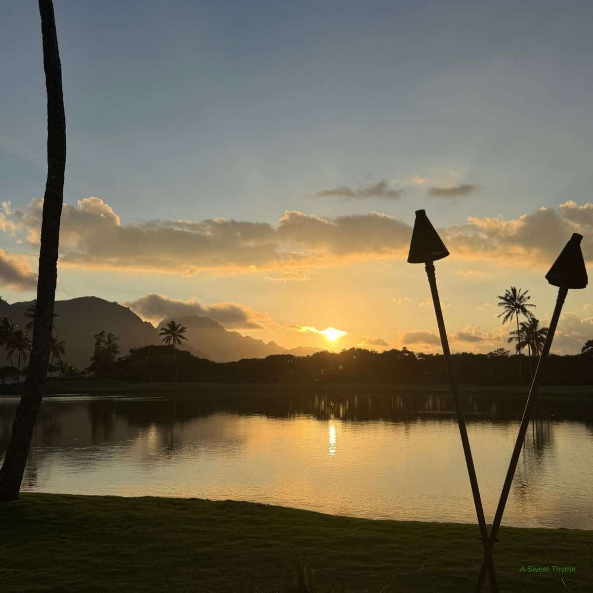Sunset over a tranquil lake with palm trees, mountains, and two tiki torches in the foreground; the sky is partly cloudy and reflects warm Sunday Thymes sunlight on the water.