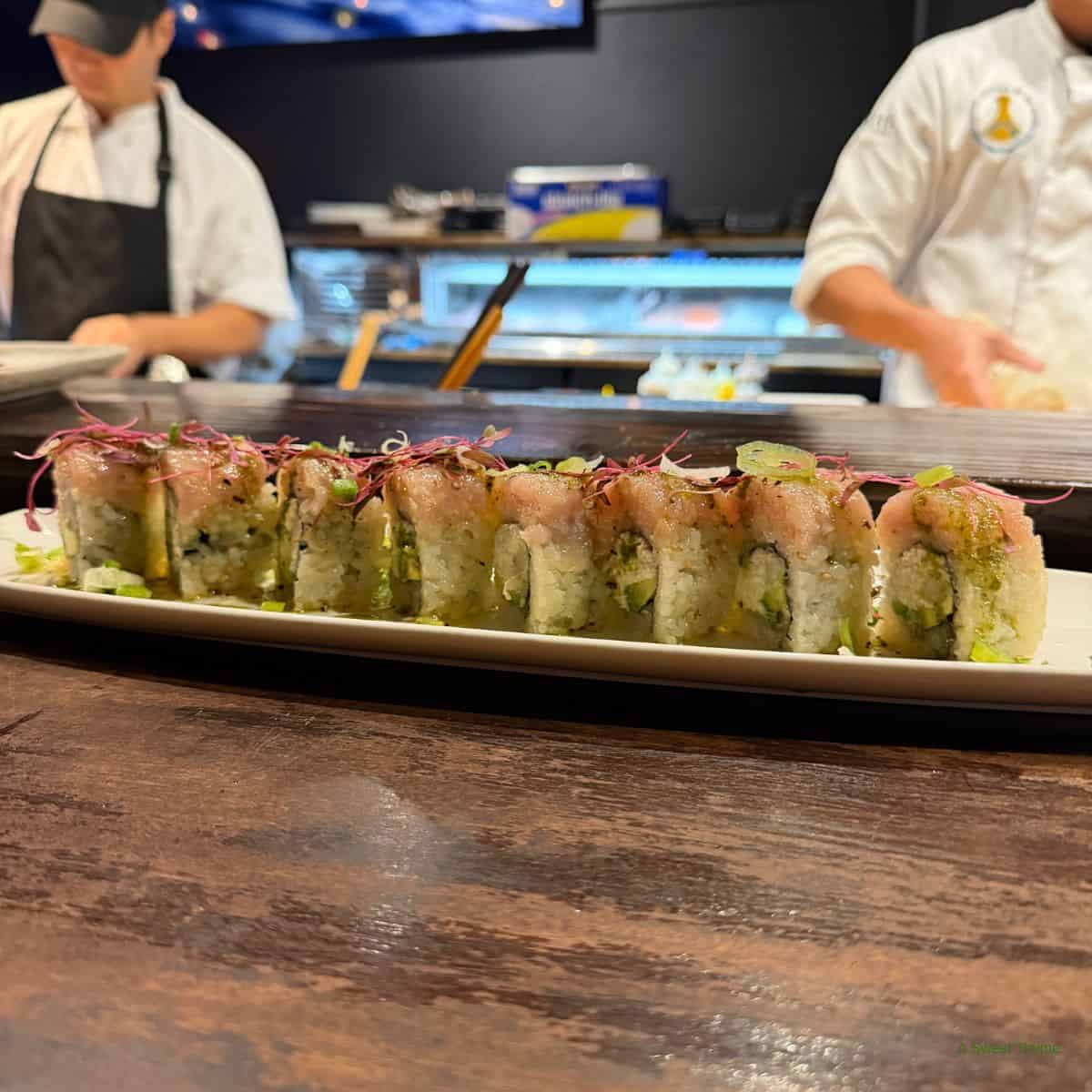 A close-up of a sushi roll topped with microgreens on a white plate at The Sunday Thymes, with two chefs preparing food in the background behind a wooden counter.