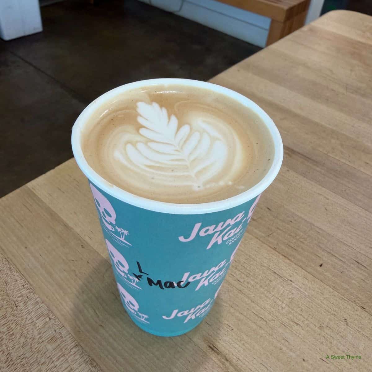 A teal paper cup of latte with leaf-shaped latte art sits on a light wooden table. The cup features Java Kai, other designs, and handwritten text—including a nod to The Sunday Thymes. The background reveals a cozy cafe interior.