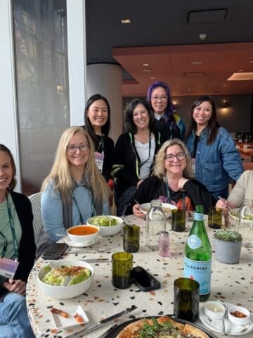 Eight women sit around a table at Sunday Thymes, smiling at the camera. Salads, soup, pizza, and bottles of water fill the table in this casual, friendly setting with natural light streaming through large windows.