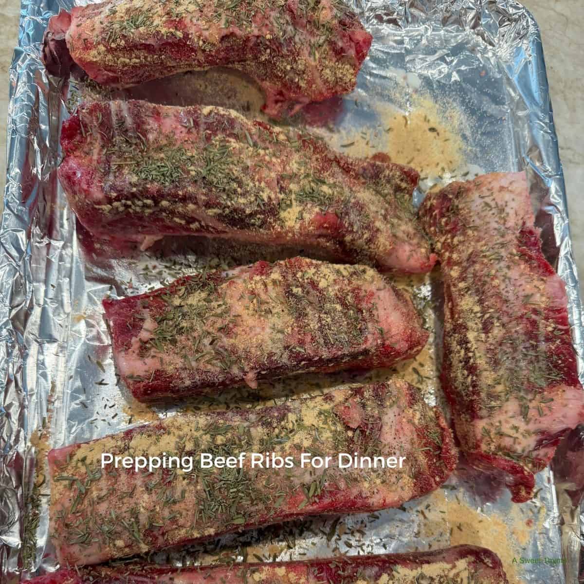 Raw beef ribs covered with herbs and seasoning are arranged on a foil-lined baking tray, ready to be cooked. The text Prepping Beef Ribs For Dinner with Sunday Thymes is overlaid on the image.