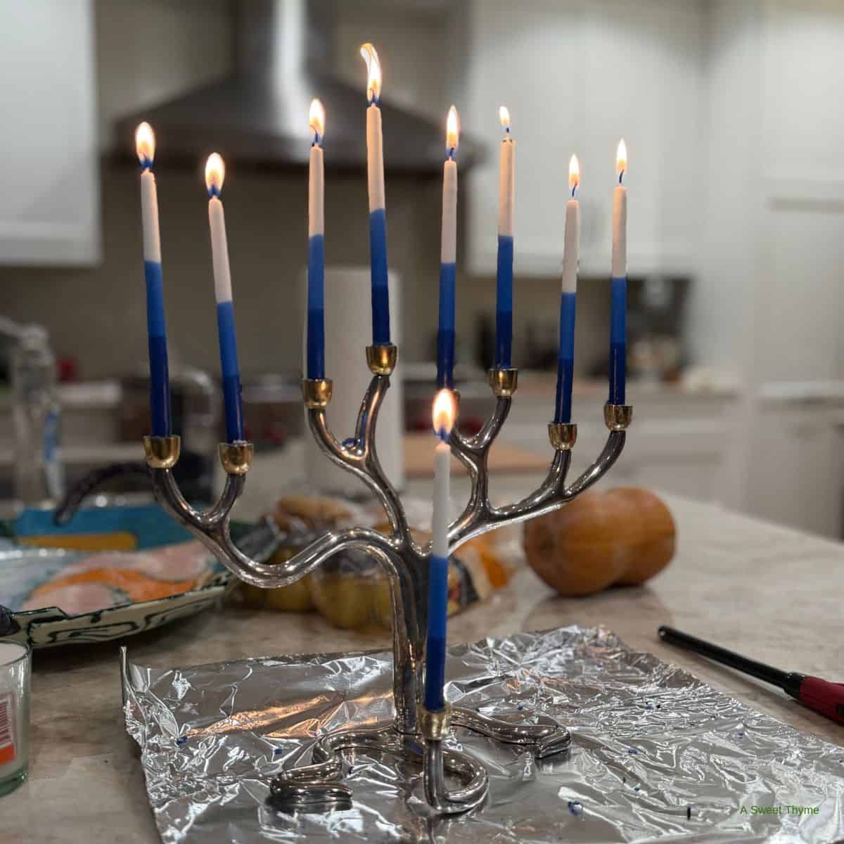 A silver menorah with nine lit blue-and-white candles sits on foil on a kitchen counter, Sunday Thymes reflected in the glow, with food and kitchen items softly blurred in the background.