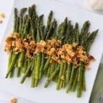 A white plate with a neatly arranged row of roasted asparagus with lemon, topped with golden brown crispy crumbs or crushed crackers.