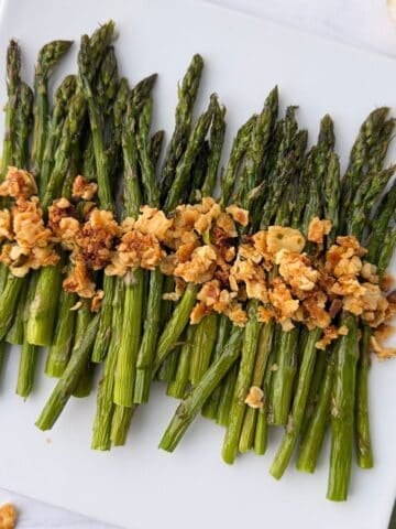 A white plate with a neatly arranged row of roasted asparagus with lemon, topped with golden brown crispy crumbs or crushed crackers.