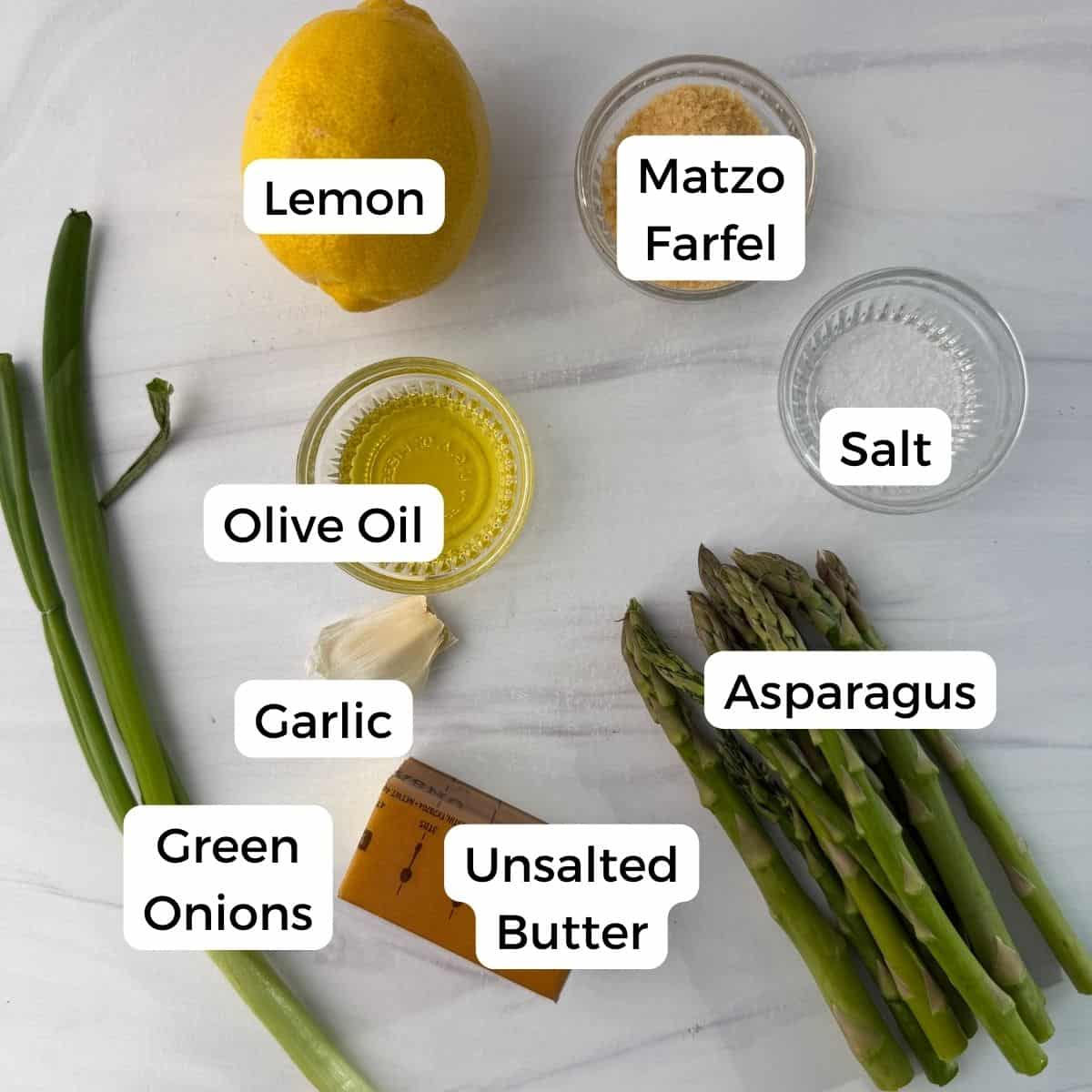 A lemon, matzo farfel, salt, green onions, olive oil, garlic, unsalted butter, and asparagus spears for roasted asparagus with lemon are arranged on a white surface, each ingredient labeled with text.