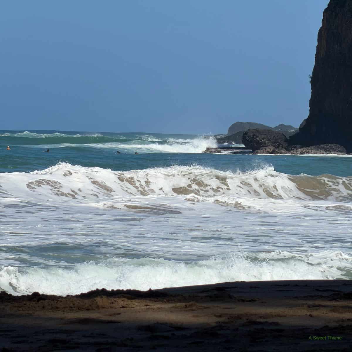 Waves crash against rocks and roll onto a sandy beach under a clear blue sky; a dark rocky cliff rises on the right side, creating a tranquil scene perfect for sunday thymes.