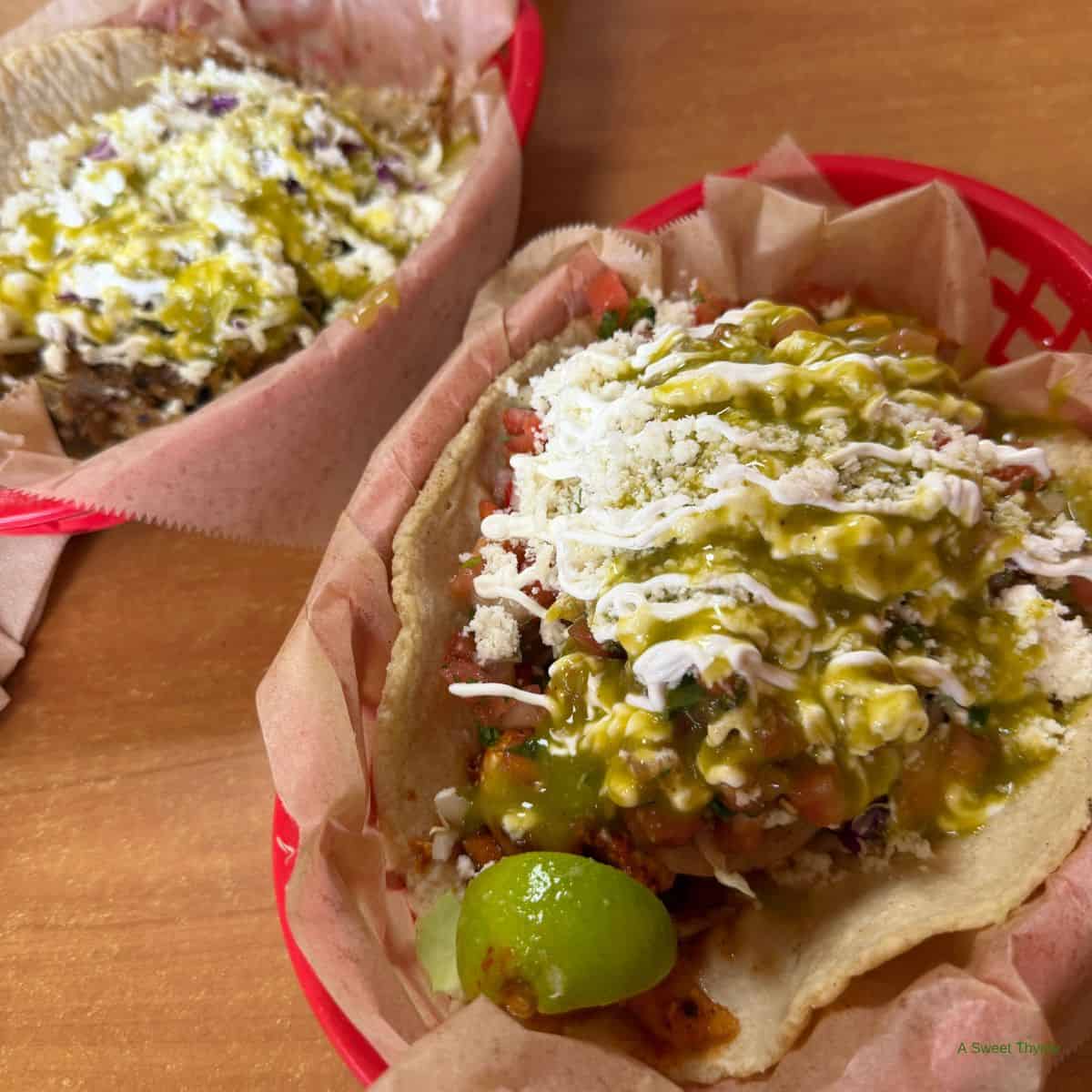 Two tacos in red baskets lined with brown paper, topped with green salsa, crumbled cheese, crema, and a lime wedge, sit on a wooden table—perfect for a relaxed Sunday Thymes meal.
