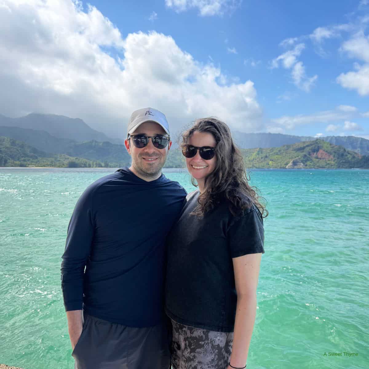 A man and woman wearing sunglasses stand and smile together in front of a turquoise ocean, with green mountains and a partly cloudy sky in the background, capturing perfect sunday thymes.