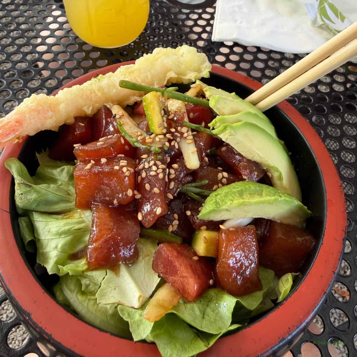 A Sunday Thymes poke bowl with marinated tuna cubes, lettuce, avocado, cucumber, green onions, and sesame seeds, topped with shrimp tempura and served in a red bowl with chopsticks on a metal table.