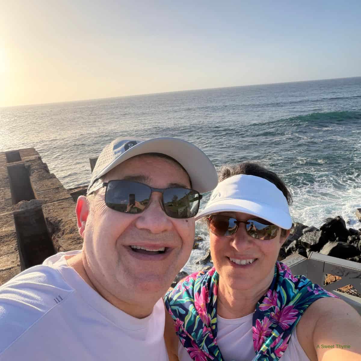 A smiling man and woman wearing Sunday Thymes sun visors and sunglasses take a selfie near an ocean pier, with waves crashing against the rocks and a clear, sunny sky in the background.