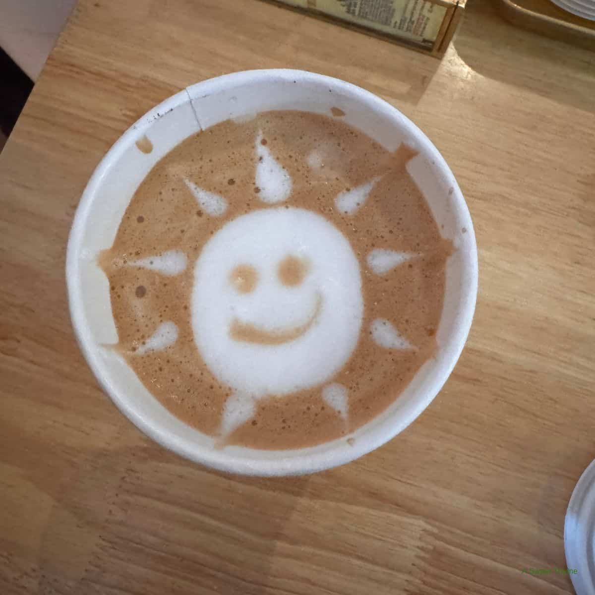 A cup of coffee with latte art showing a smiling sun with rays, set on a wooden table—perfect for slow mornings and Sunday Thymes.