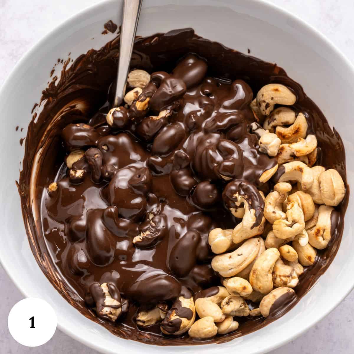 A white bowl filled with partially mixed melted chocolate and whole cashews resembles the start of a trifle with chocolate. A metal spoon rests in the glossy, rich mixture. A small white circle labeled 1 appears in the bottom left corner.
