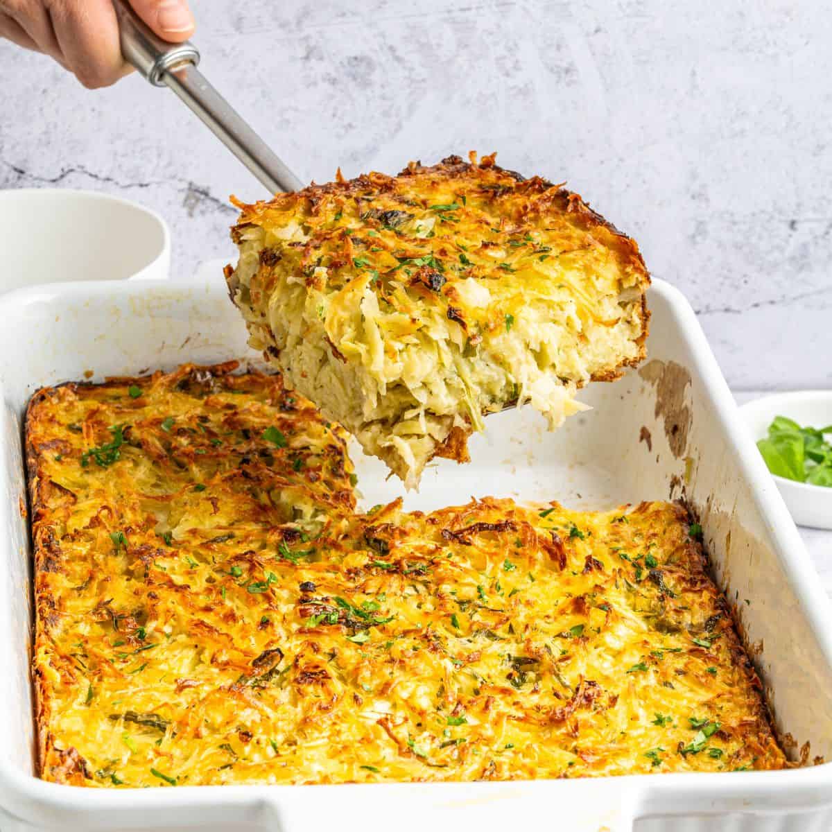 A hand lifts a large serving of golden-brown potato kugel casserole from a white rectangular dish. The dish boasts a crispy top, with shredded potatoes and green herbs peeking through.