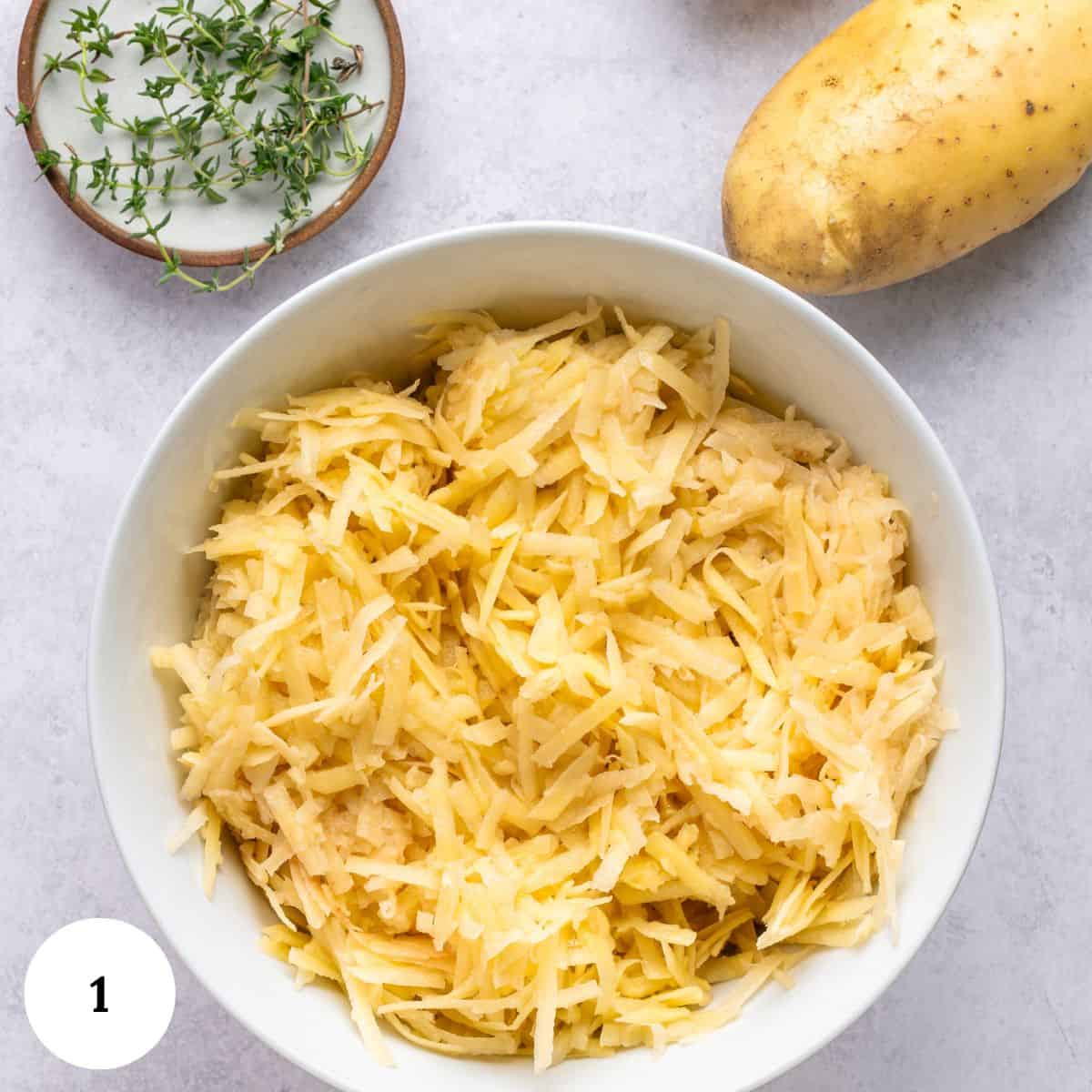 A white bowl filled with shredded potatoes for potato kugel sits on a light countertop, next to a whole potato, a small dish of fresh thyme, and a round label with the number 1.