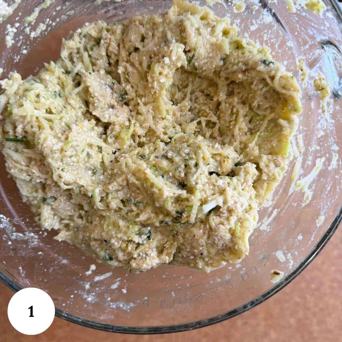 A glass bowl filled with a thick, textured zucchini kugel mixture of shredded vegetables and flour, resembling a batter or dough, with visible green herbs and a slightly lumpy consistency.