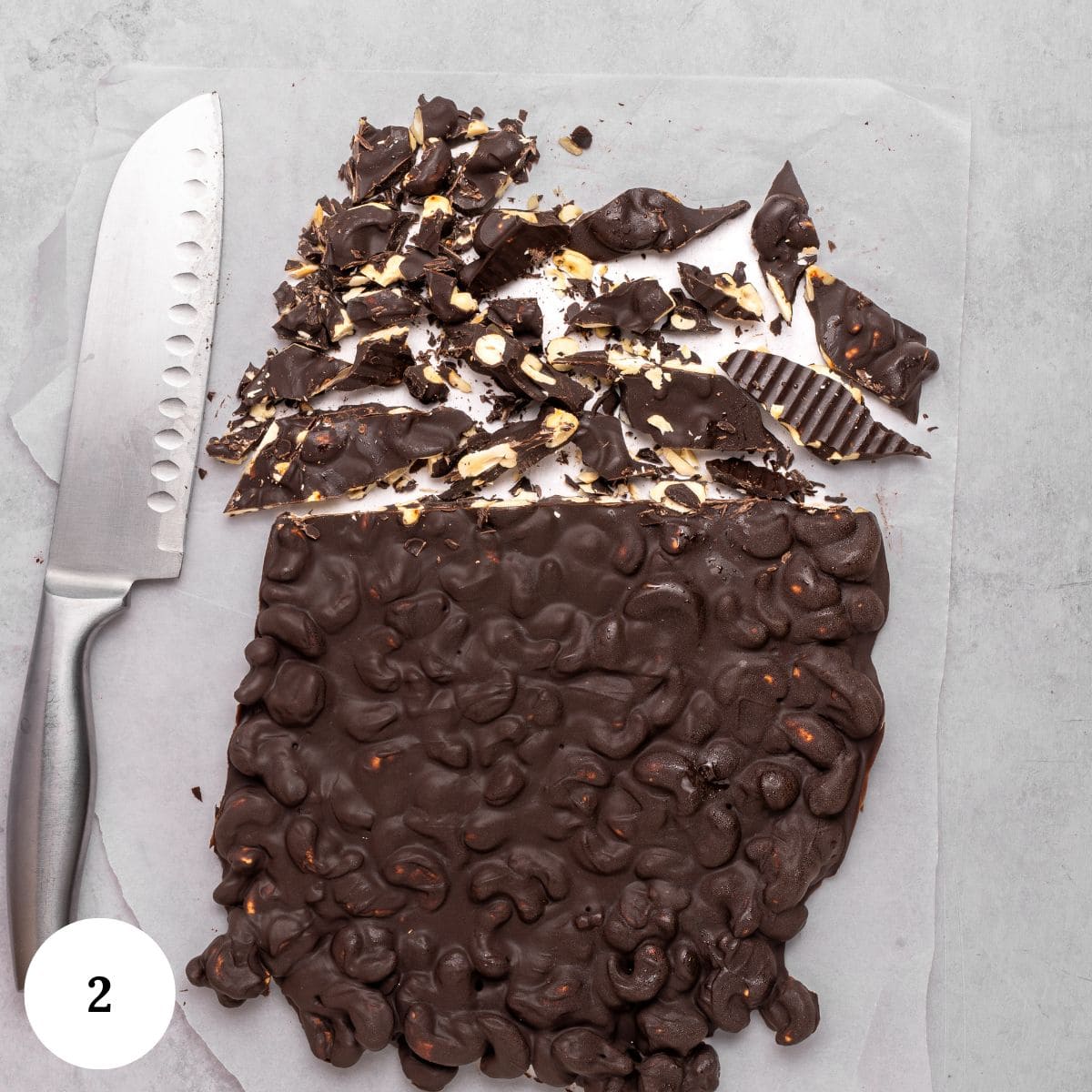 A large block of nut-studded dark chocolate, perfect for a trifle with chocolate, is partially chopped into pieces on parchment paper. A knife rests beside it on a light gray surface, and a white circle with the number 2 appears in the bottom left corner.