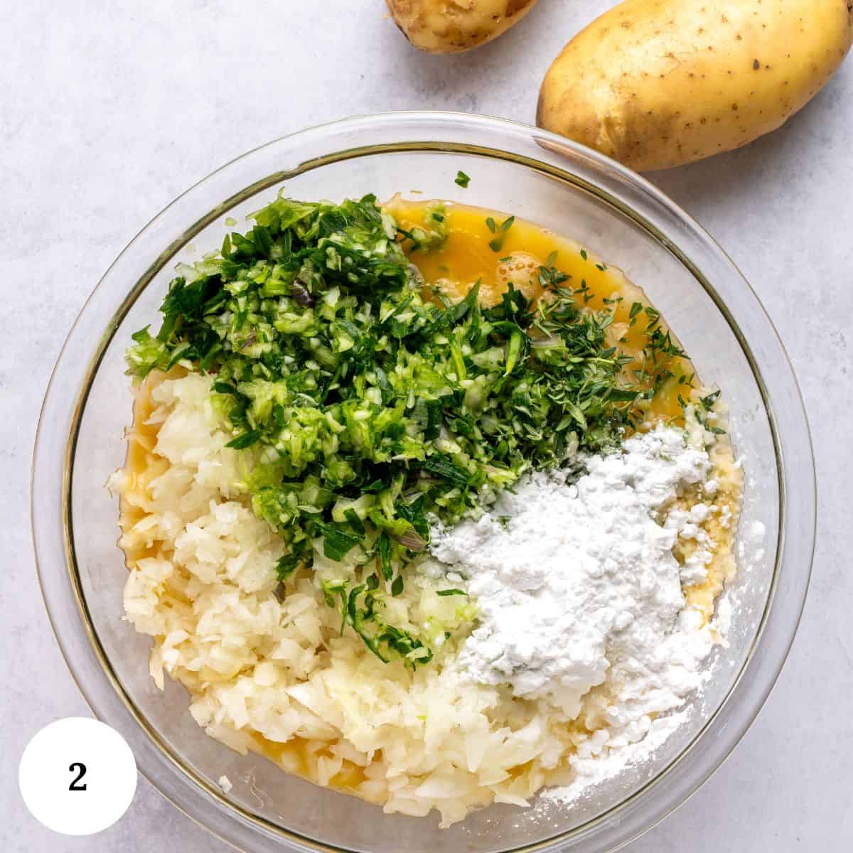 A glass bowl containing grated potatoes, chopped herbs, flour, and eggs sits on a light surface-ingredients ready for potato kugel. Two whole potatoes are nearby. The number 2 appears in a white circle in the bottom left corner.