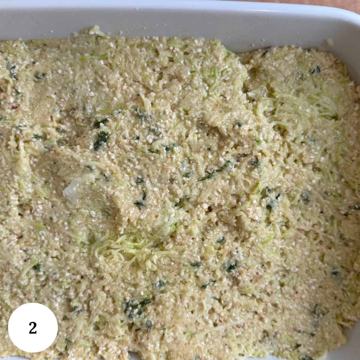 A white baking dish holds an uncooked, textured mixture of shredded vegetables and herbs, resembling a zucchini kugel with cheese and seasonings. A white circle with the number 2 appears in the lower left corner.