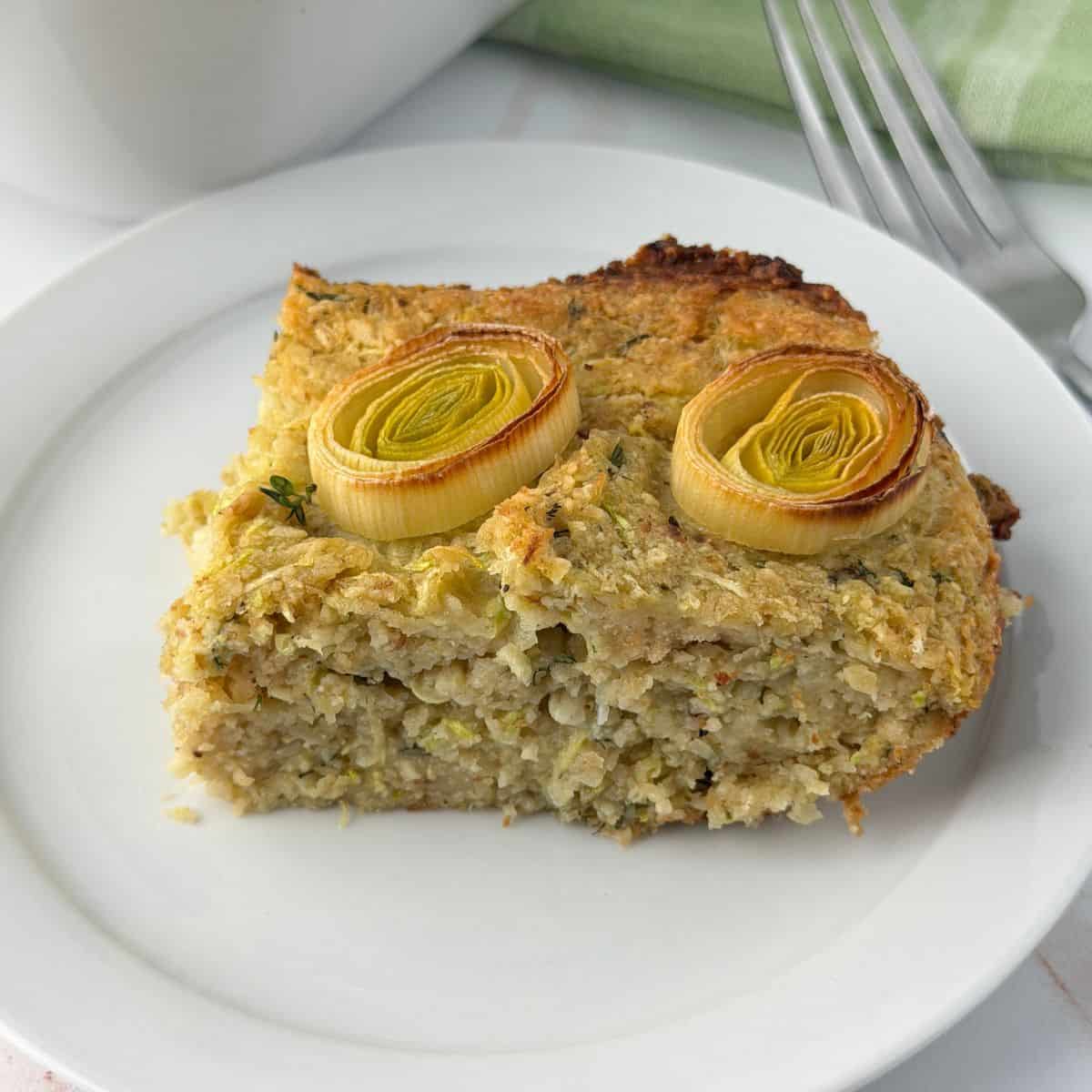 A slice of savory zucchini kugel casserole, topped with two round slices of cooked leek, is served on a white plate with a fork and a green napkin in the background.