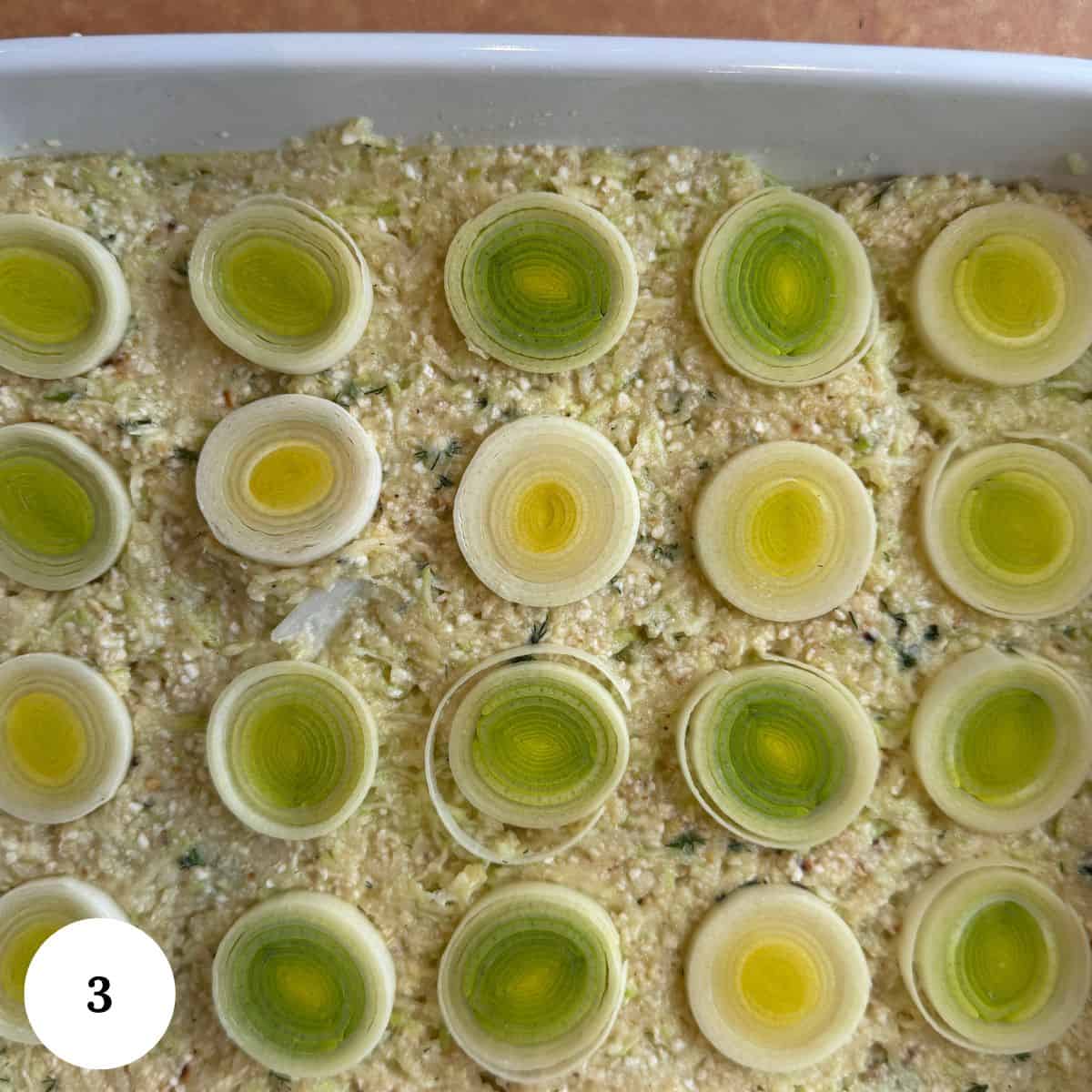 A baking dish filled with a textured, creamy zucchini kugel mixture is topped with evenly spaced slices of leek, displaying concentric green and yellow rings. A white circle labeled 3 is in the bottom left corner.
