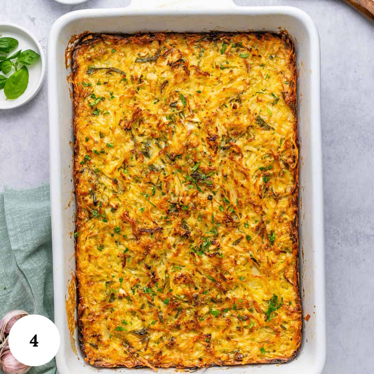Golden-brown baked potato kugel casserole in a white rectangular dish, garnished with fresh herbs. The dish is set on a light gray surface with garlic, basil, and a green cloth nearby.