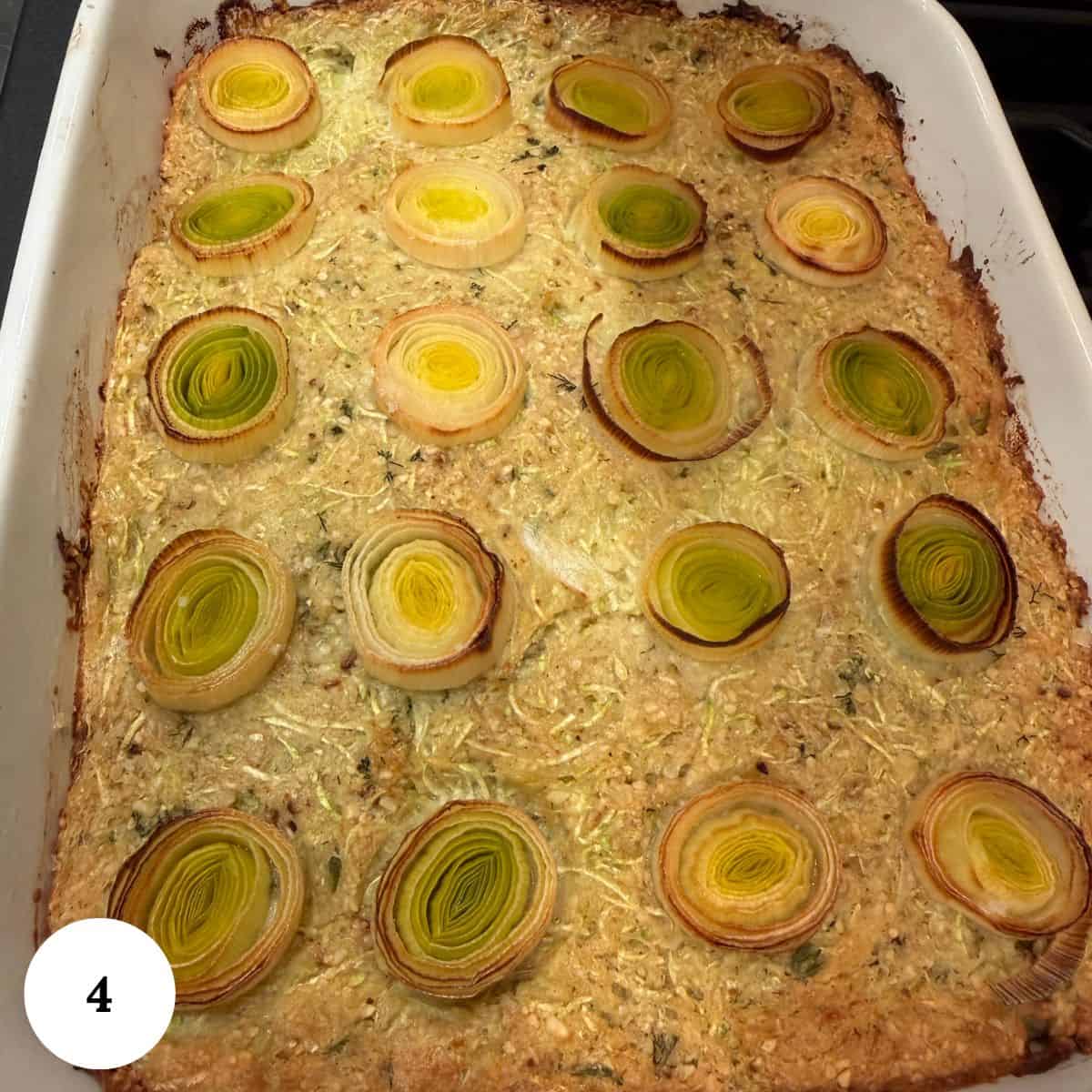 A baked zucchini kugel casserole topped with evenly spaced, cross-sectional slices of leeks arranged in rows on a golden, textured surface in a rectangular white dish. A small white circle with the number 4 appears in the lower left corner.