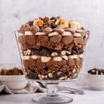 A glass trifle bowl filled with layers of chocolate mousse, bananas, chopped nuts, and chocolate pieces—a decadent trifle with chocolate—topped with more bananas and chocolate crumbles. Bowls of extra toppings sit in the background.