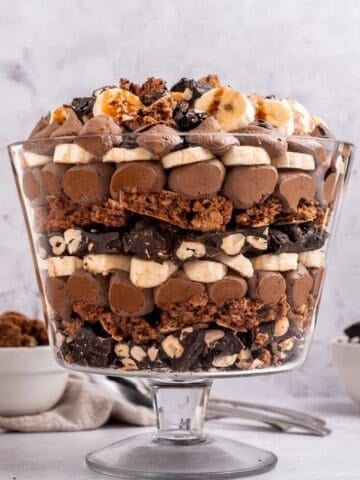 A glass trifle bowl filled with layers of chocolate mousse, bananas, chopped nuts, and chocolate pieces-a decadent trifle with chocolate-topped with more bananas and chocolate crumbles. Bowls of extra toppings sit in the background.
