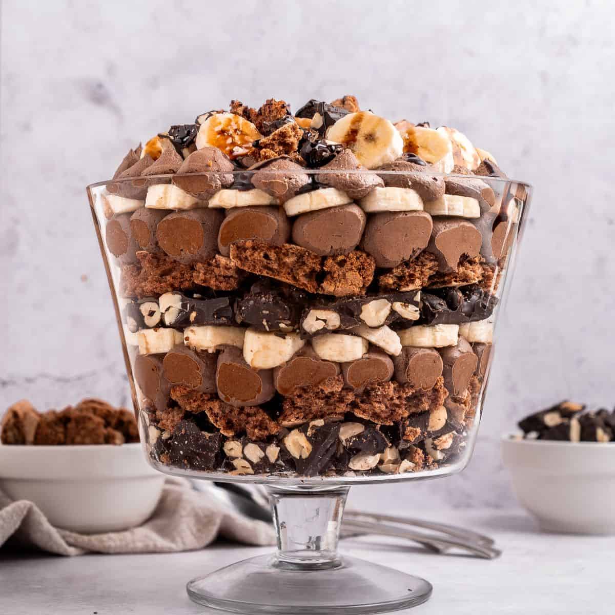 A glass trifle bowl filled with layers of chocolate mousse, bananas, chopped nuts, and chocolate pieces-a decadent trifle with chocolate-topped with more bananas and chocolate crumbles. Bowls of extra toppings sit in the background.