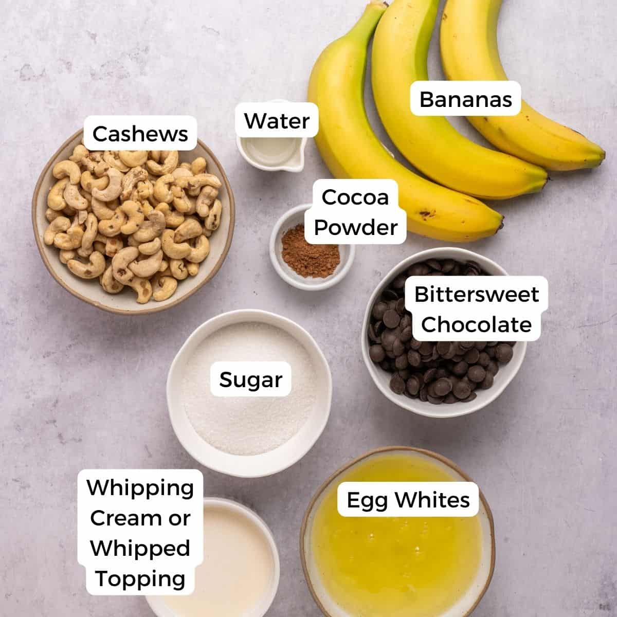 Top-down view of bowls containing cashews, cocoa powder, bittersweet chocolate, sugar, egg whites, and whipping cream—ingredients perfect for a trifle with chocolate—alongside whole bananas and a small bowl of water, all labeled with text.