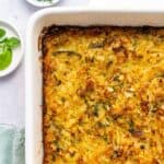 A close-up of a golden-brown potato kugel casserole with a crispy top in a white dish, surrounded by small plates with fresh greens and a green cloth, on a light-colored surface.