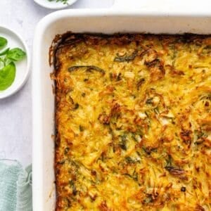 A close-up of a golden-brown potato kugel casserole with a crispy top in a white dish, surrounded by small plates with fresh greens and a green cloth, on a light-colored surface.