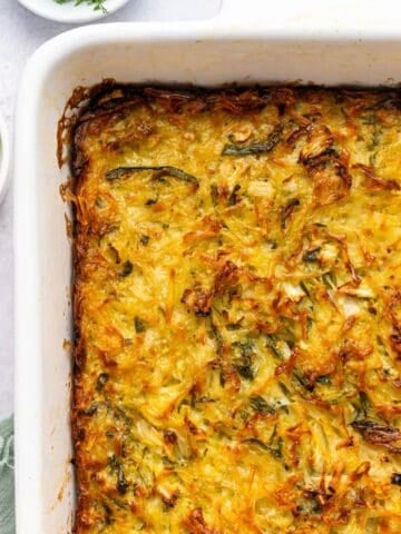 A close-up of a golden-brown potato kugel casserole with a crispy top in a white dish, surrounded by small plates with fresh greens and a green cloth, on a light-colored surface.