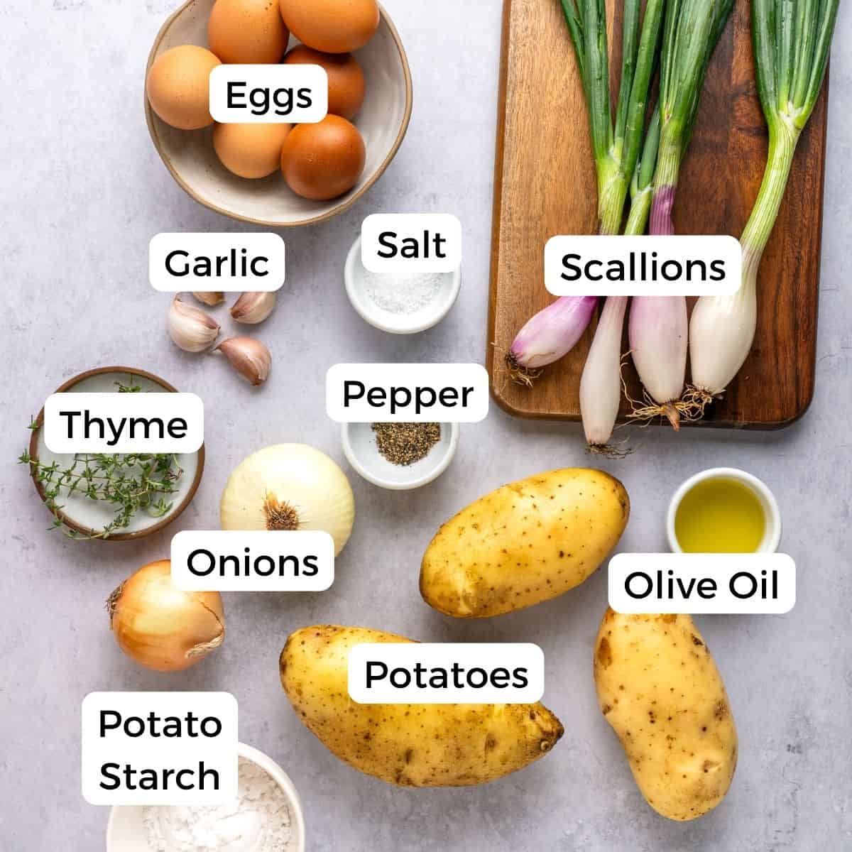 A variety of labeled ingredients for potato kugel are arranged on a table, including eggs, garlic, thyme, onions, potato starch, potatoes, salt, pepper, scallions, and a small bowl of olive oil.