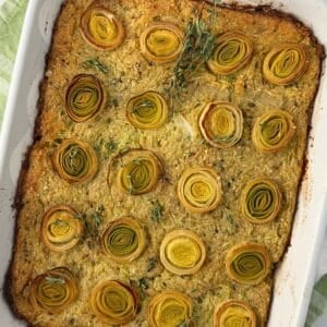 A baked casserole reminiscent of zucchini kugel, topped with neatly arranged rounds of leeks and garnished with fresh thyme in a white rectangular dish. The edges are browned and the vegetables are beautifully golden.