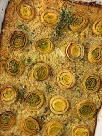 A baked casserole reminiscent of zucchini kugel, topped with neatly arranged rounds of leeks and garnished with fresh thyme in a white rectangular dish. The edges are browned and the vegetables are beautifully golden.