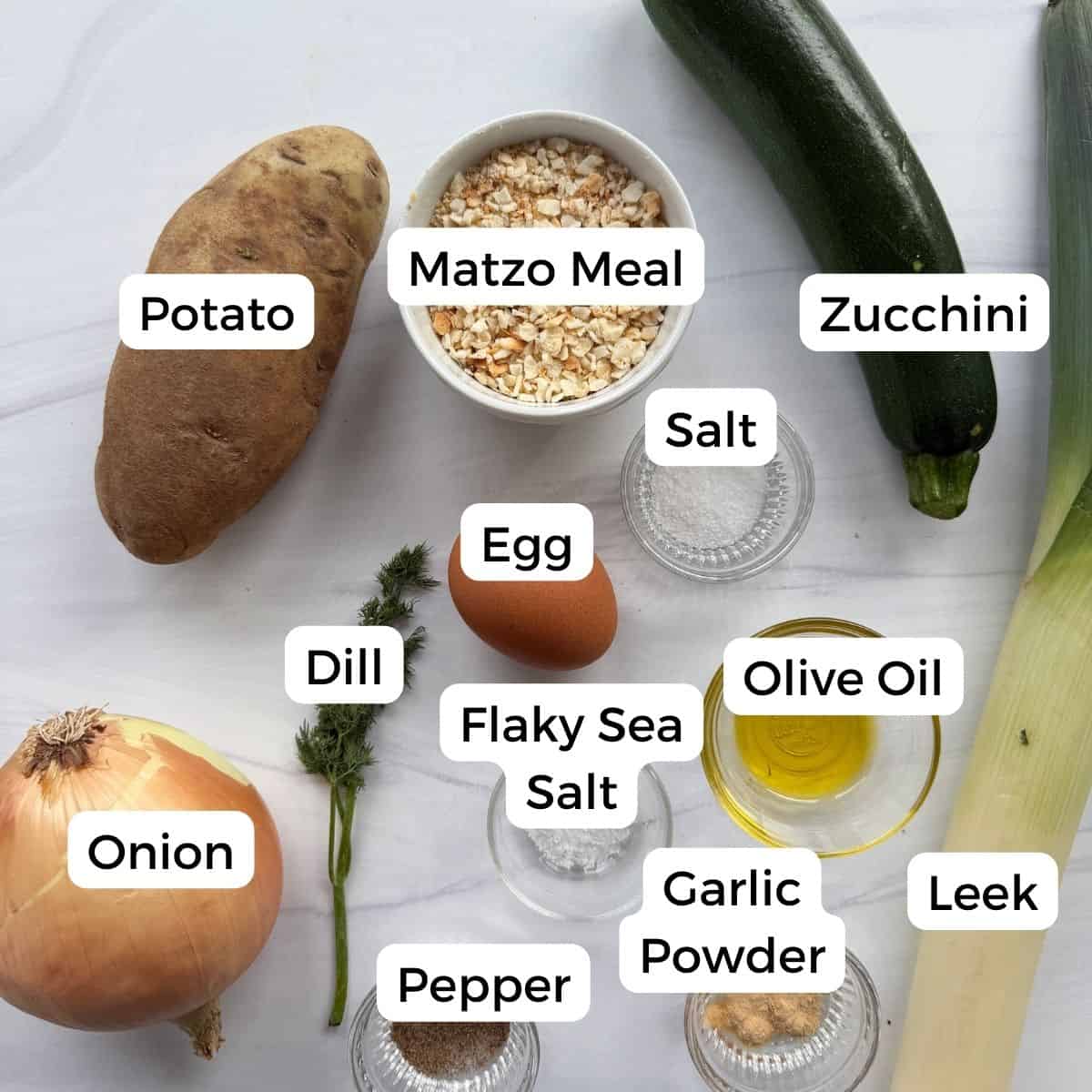 A top view of labeled ingredients for zucchini kugel: potato, zucchini, leek, onion, matzo meal, egg, dill, salt, flaky sea salt, pepper, garlic powder, and olive oil on a white surface.