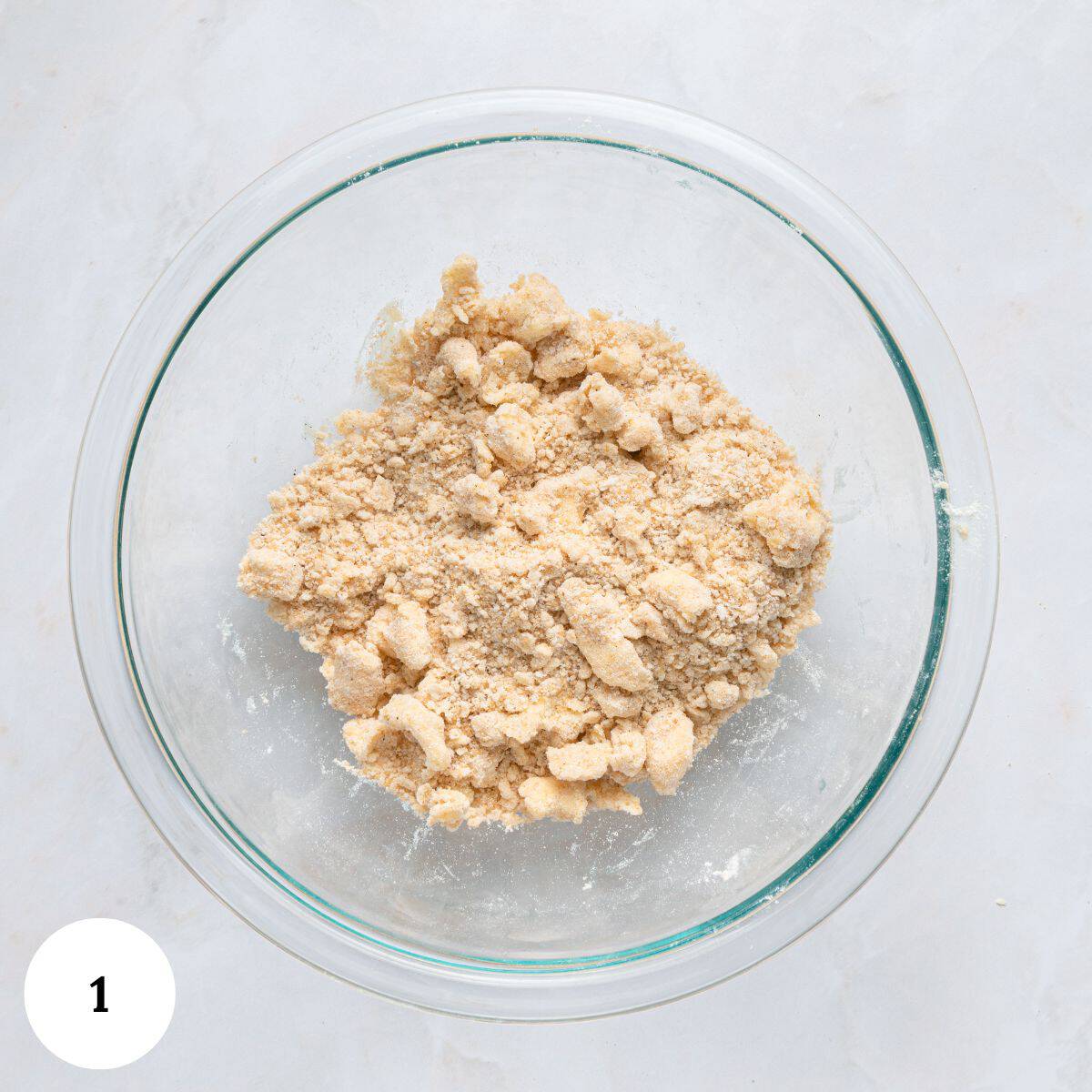 A clear glass bowl filled with crumbly dough mixture for blueberry yogurt muffins sits on a white marble surface. A white circle with the number 1 is in the bottom left corner of the image.