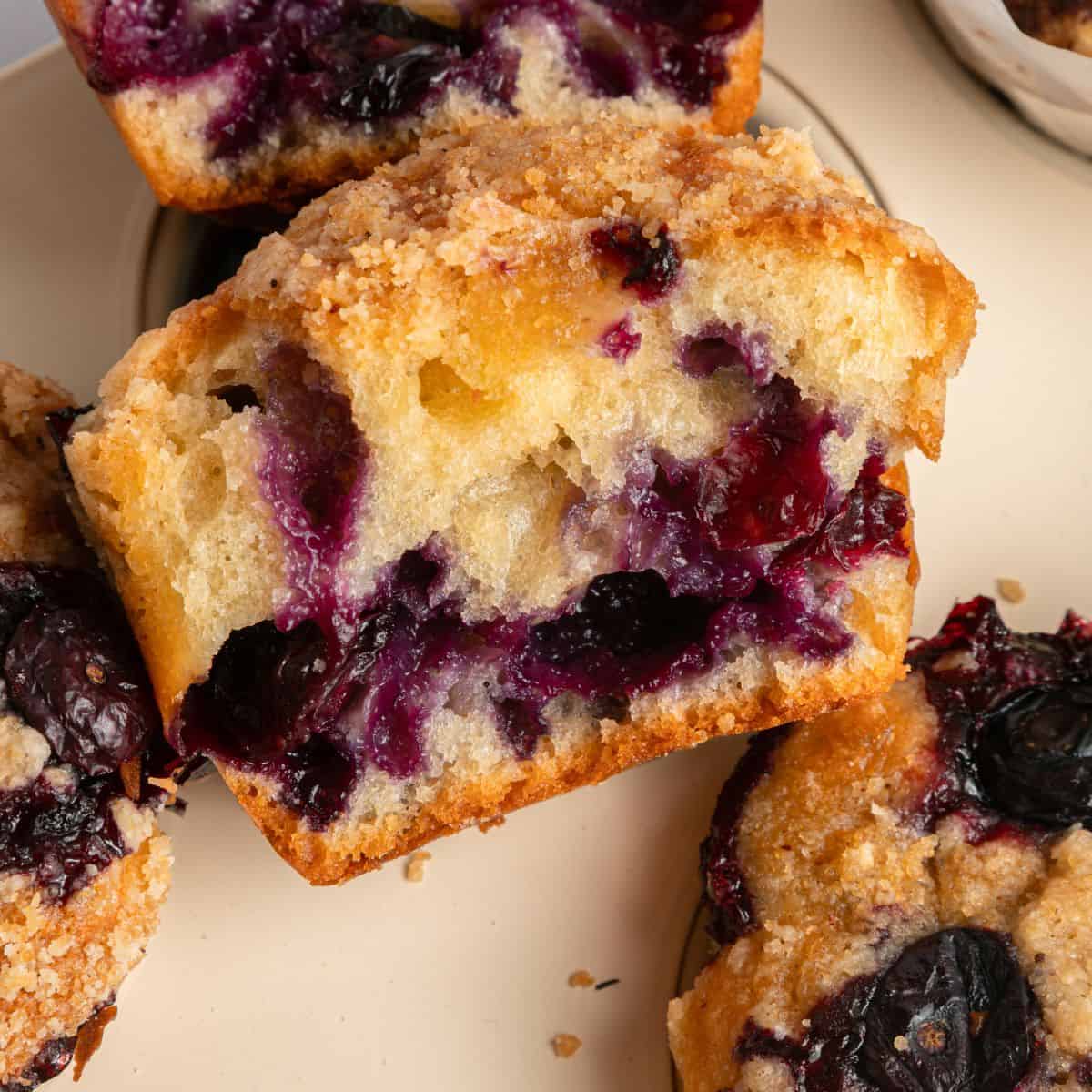 A close-up of sliced blueberry yogurt muffins with a crumbly topping, showing juicy blueberries baked inside the moist, golden muffin. The muffins are stacked on a beige plate.
