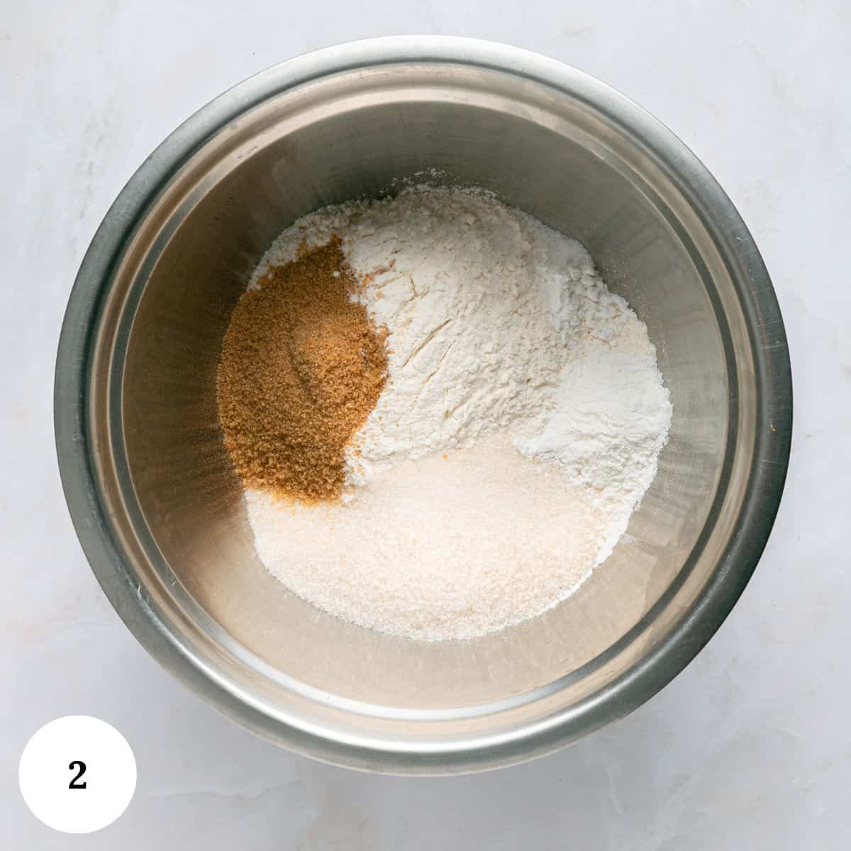 A metal mixing bowl containing separate piles of brown sugar, white flour, and granulated sugar on a light marble surface-ingredients ready for blueberry yogurt muffins. A white circle with the number 2 is in the bottom left corner.